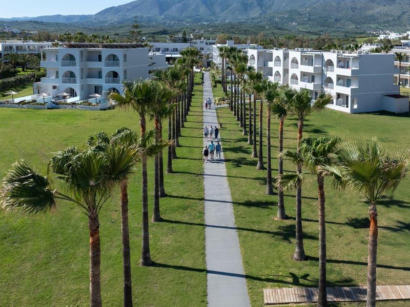 Blick auf einen langen Gehweg mit Palmen zwischen zwei weißen Hotelgebäuden und Bergen im Hintergrund.