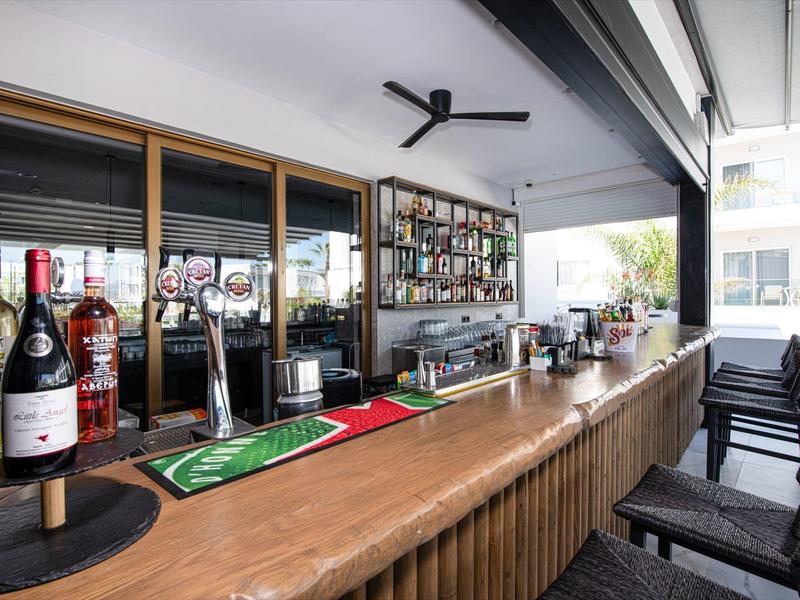 Moderner Barbereich mit Holztheke, Weinflaschen und Sitzstühlen in einem hellen Raum.