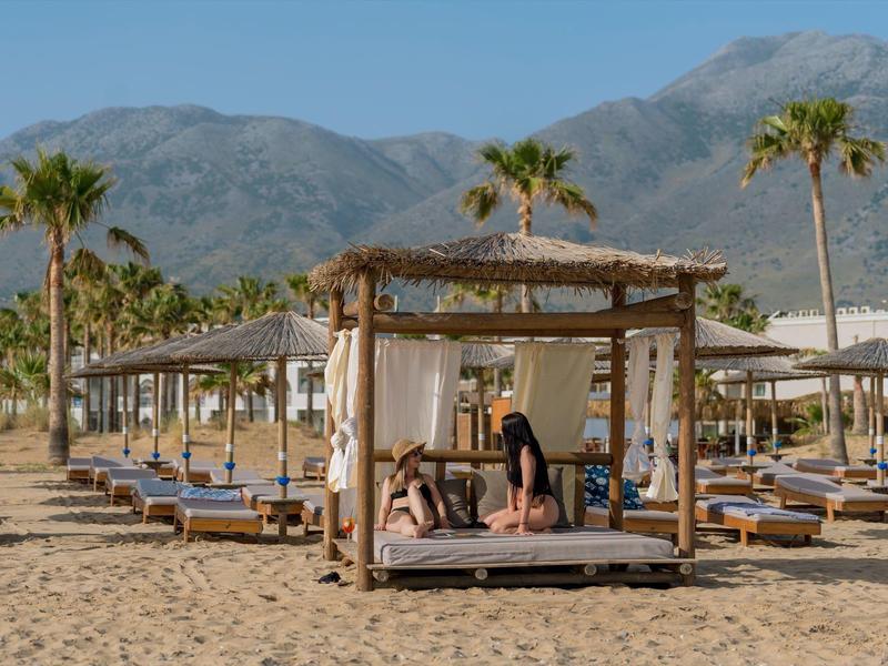 Strand mit Holzpavillon, Liegen und Palmen vor Bergkulisse bei sonnigem Wetter.