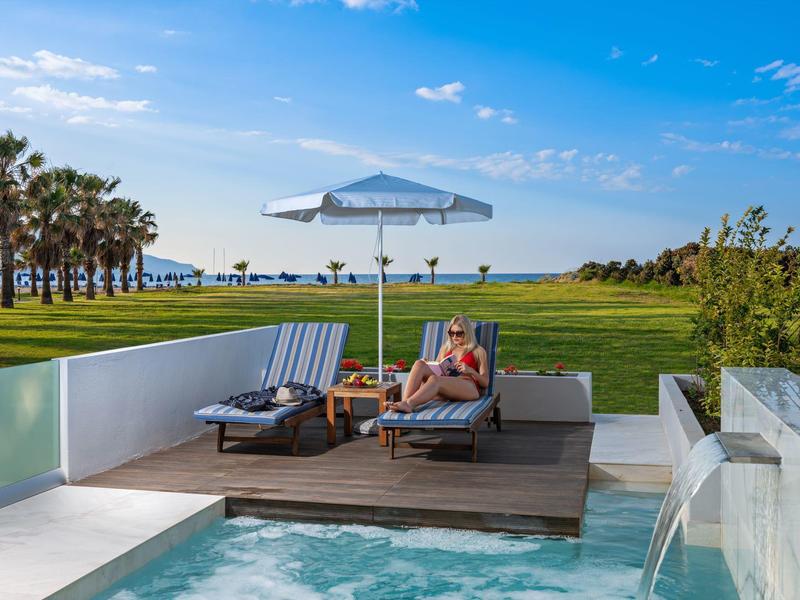 Luxuriöse Poolterrasse mit Liegestühlen und Sonnenschirm vor einem grünen Rasen und Blick aufs Meer.