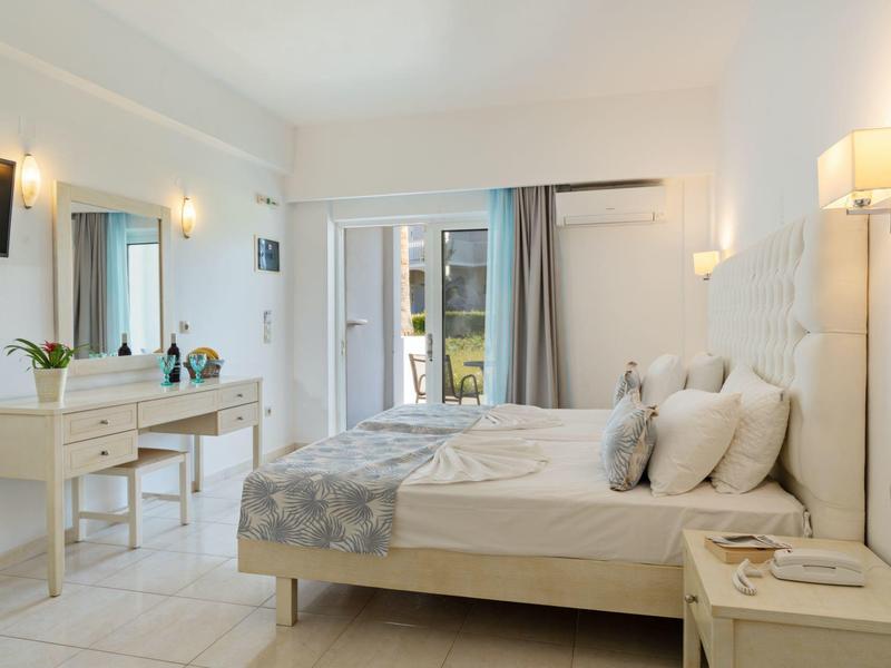 Helles Schlafzimmer mit weißem Bett, Sideboard, TV, Lampe und Terrassentür mit Vorhängen.