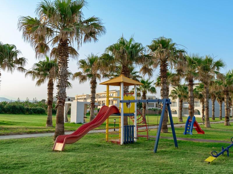 Kinderspielplatz mit Klettergerüst, Rutsche, Schaukel und Palmen im Hintergrund bei blauem Himmel.