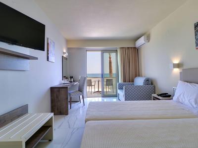 Jasny pokój hotelowy z dwoma łóżkami, biurkiem, telewizorem i balkonem z widokiem na morze.