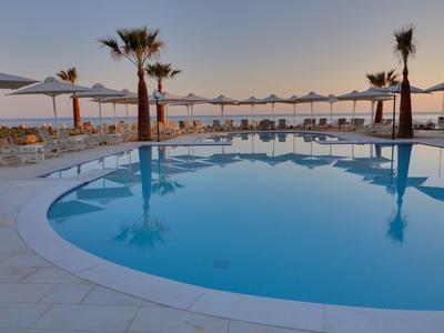 Piscina rotonda con lettini, ombrelloni e palme al tramonto vicino al mare.