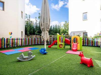 Plac zabaw dla dzieci z zjeżdżalniami, huśtawką i kolorowym ogrodzeniem między białymi budynkami.