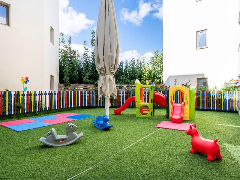 Plac zabaw dla dzieci z zjeżdżalniami, huśtawką i kolorowym ogrodzeniem między białymi budynkami.