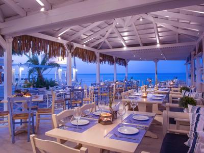 Open seaside restaurant with set tables and wooden furniture under a thatched roof.