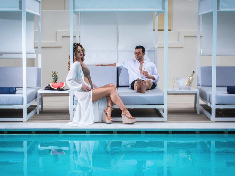 Two people relax on a poolside daybed next to a clear blue pool.