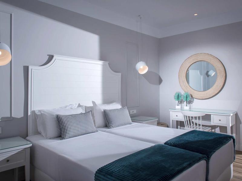 Bright hotel room with two beds, white furniture, and blue blankets at the foot of the beds.