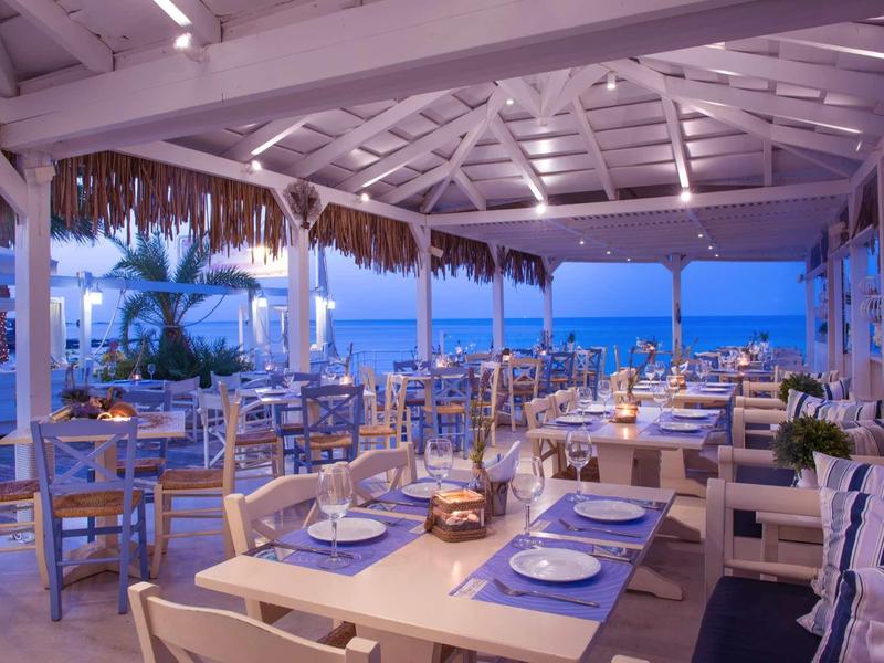 Open seaside restaurant with set tables and wooden furniture under a thatched roof.