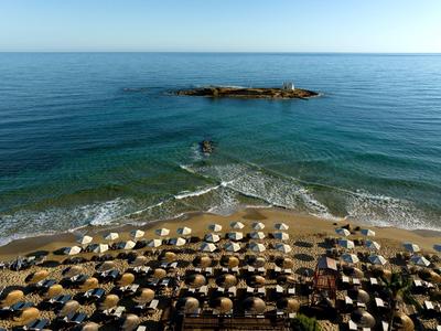 Pláž s řadami slunečníků a malou skalní formací v čistém moři.