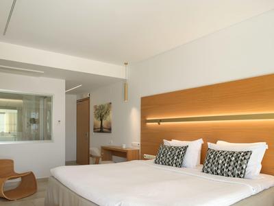 Moderní hotelový pokoj s velkou postelí, dřevěnými prvky a stolem vedle skleněné stěny.