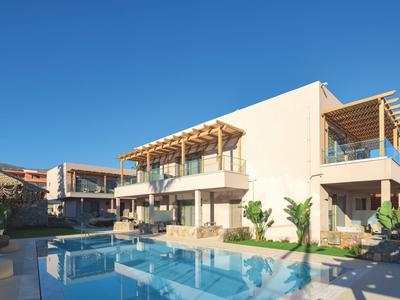 Moderní hotel s venkovním bazénem a kvetoucími rostlinami pod jasně modrou oblohou.