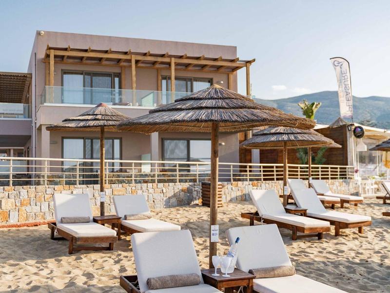 Plážová lehátka a slunečníky před moderním hotelem pod jasnou oblohou.