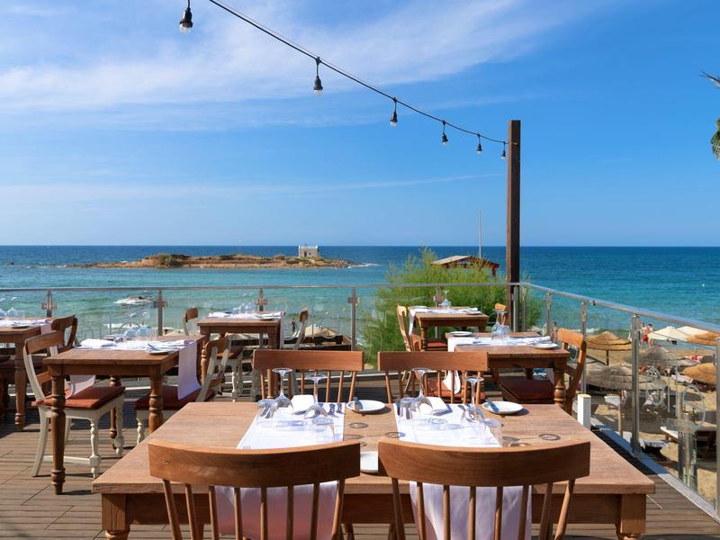 Venkovní restaurace s dřevěnými stoly s výhledem na moře pod jasnou oblohou.