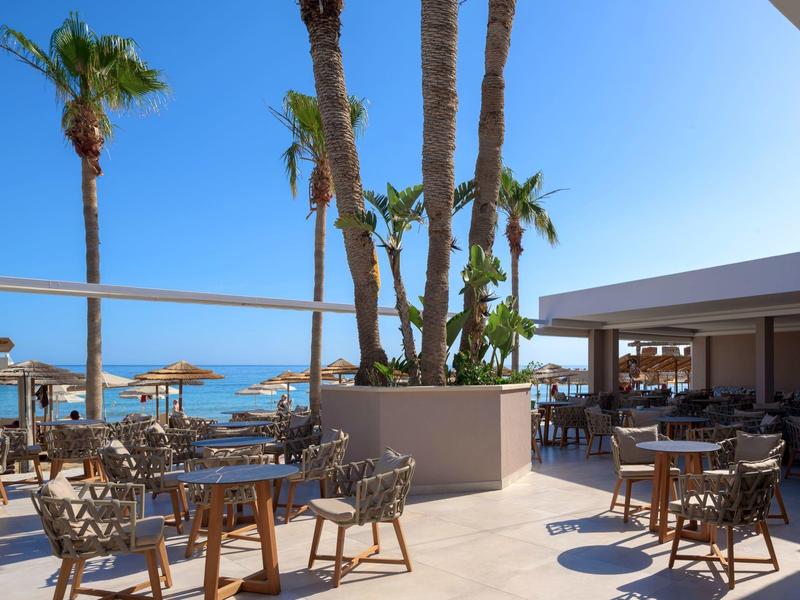 Otevřená venkovní terasa hotelu s stoly, židlemi a palmami pod jasnou oblohou.