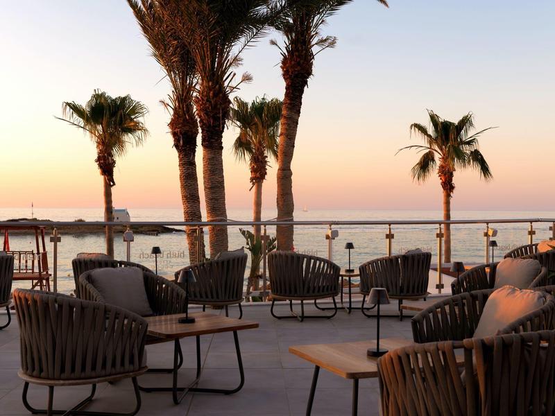 Venkovní lounge s židlemi a stoly, palmy a moře na pozadí západu slunce.