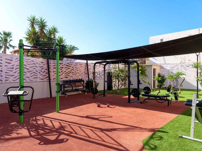 Moderní venkovní fitness prostor se zařízeními na červené podlaze vedle trávy a bílých zdí.