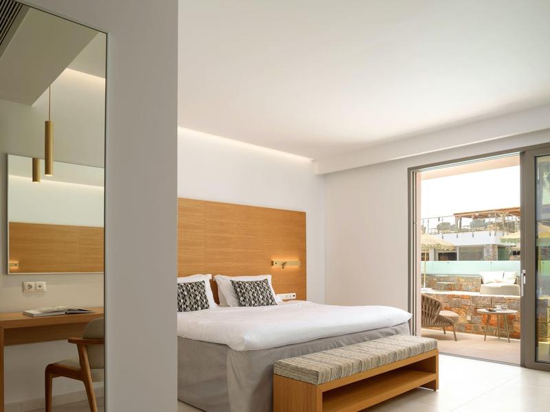 Světý hotelový pokoj s manželskou postelí, dřevěnými detaily a balkonem s výhledem na budovy.