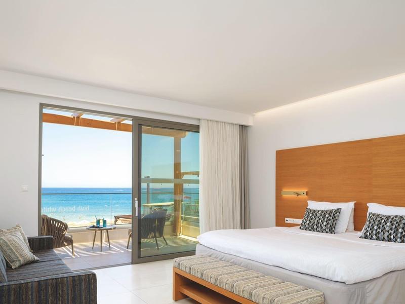 Moderní hotelový pokoj s manželskou postelí, pohovkou a výhledem na moře přes velká skleněná okna.