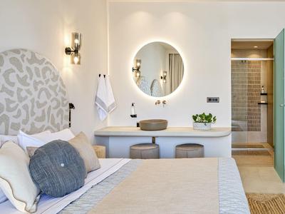 Helles Schlafzimmer mit rundem Spiegel, grauem Kopfteil, zwei Hockern und modernem Bad im Hintergrund.