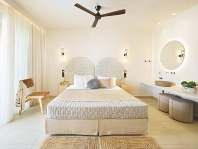 Helles Schlafzimmer mit großem Bett, rundem Spiegel, Deckenventilator und Holzstuhl am Fenster.