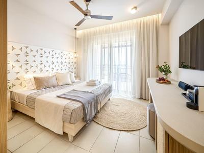 Helles Schlafzimmer mit Doppelbett, Beistelltisch, Vorhängen, Deckenventilator und großer Wandfernseher.