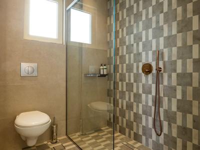 Moderner hellgrauer Duschbereich mit Glaswand, braunem Regenbrausekopf und WC an heller Wand.