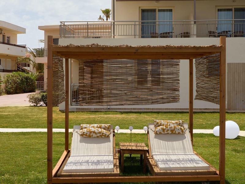 Two lounge chairs under a wooden gazebo on green grass in front of a modern building.