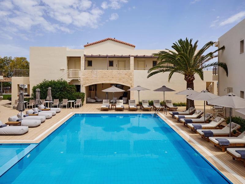 Modern outdoor pool with lounge chairs and umbrellas in front of a light hotel building.