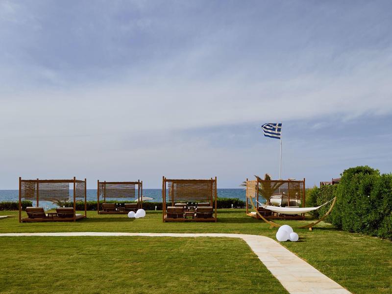 Green lawn with loungers, sunshades, and Greek flag by the sea under cloudy sky.