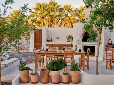 Przytulna przestrzeń na zewnątrz z drewnianymi stołami, krzesłami i kominkiem, otoczona roślinami i palmami.