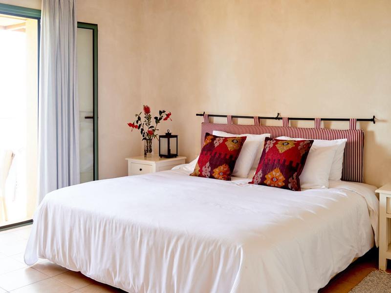 Camera luminosa con grande letto, biancheria bianca e cuscini colorati vicino al balcone.