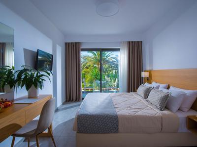 Helles Hotelzimmer mit großem Bett, Schreibtisch, Fernseher und Blick auf grüne Palmen.