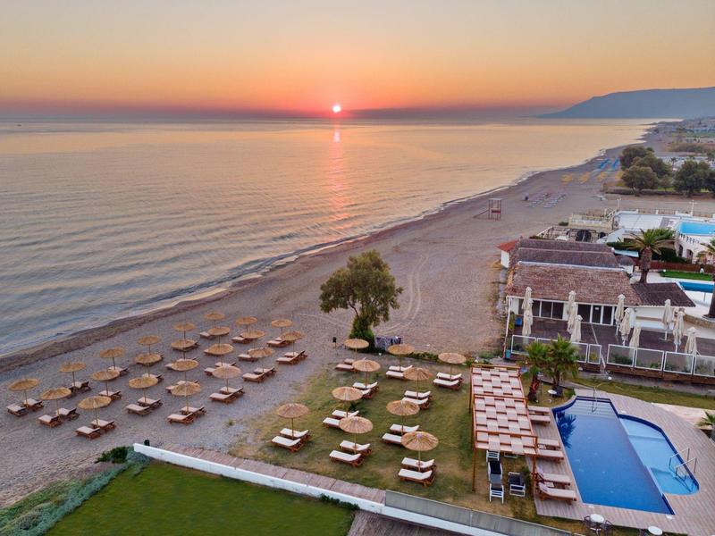 Zonsondergang boven een rustig strand met ligstoelen en een zwembad naast hotelgebouwen.