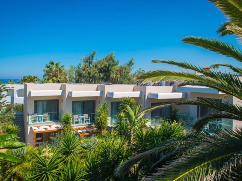 Moderne witte hotelkamers met balkons omgeven door weelderige tropische planten en een heldere blauwe lucht.