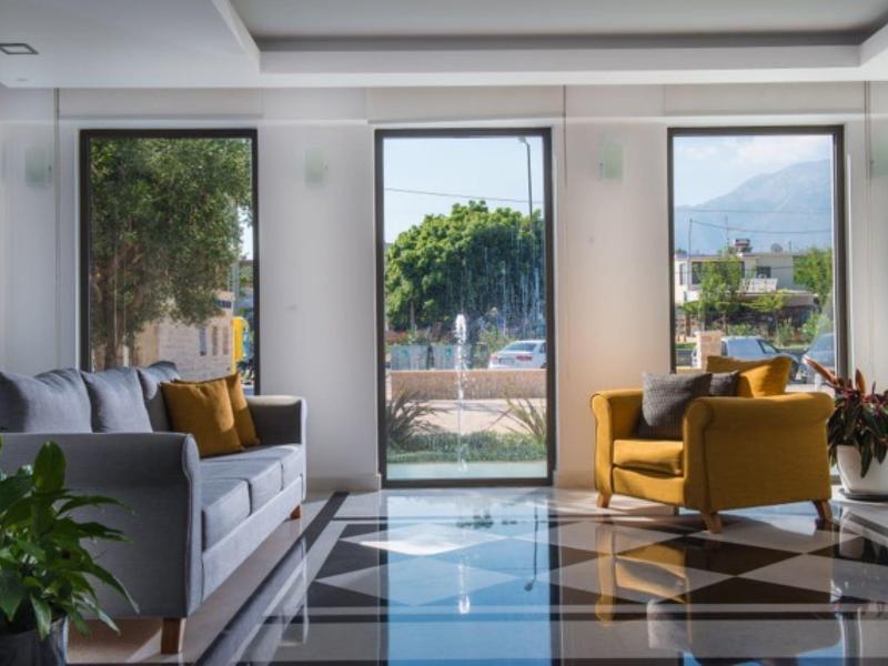 Lichte hotel lounge met grijze bank, mosterdkleurige fauteuils, grote ramen en betegelde vloer.