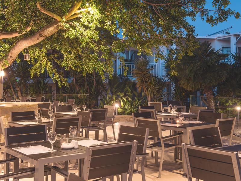 Gezellige buitenruimte van een restaurant met tafels en stoelen onder bomen verlicht bij schemering.