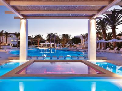 Evening view of illuminated hotel pool with hot tub and palm trees under pergola.