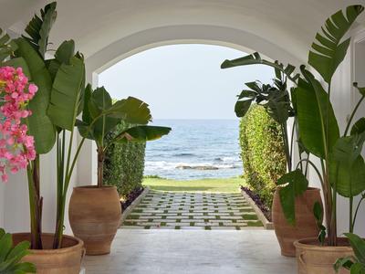 View through a white archway to the beach, with green plants and pots on both sides in the foreground.