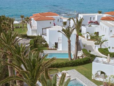 White villas with red roofs by the coast with palm trees and a pool.