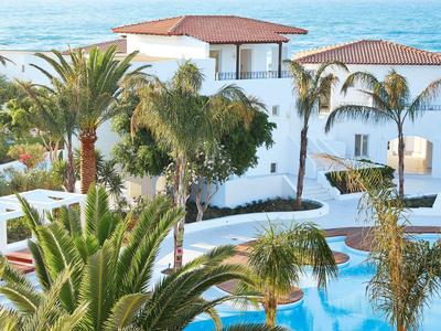 White hotel with red roofs surrounded by palm trees and a blue outdoor pool.