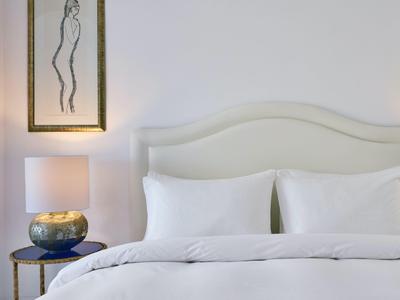 Minimalistický hotelový pokoj s bílou postelí, nočním stolkem s lampou a jednoduchým obrazem na zdi.