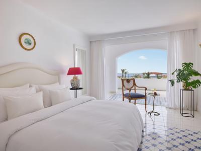 Heller Hotelzimmer mit großem Bett, Fliesenboden und Blick auf Terrasse mit Meer.