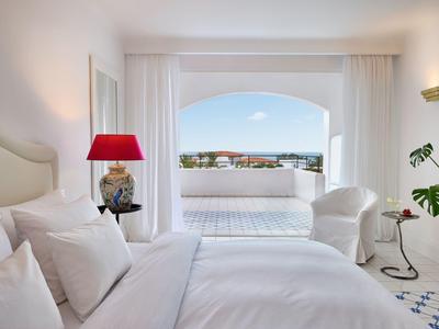 Helles Hotelzimmer mit weißer Bettwäsche, rotem Lampenschirm und Balkon mit Aussicht auf eine Stadt.