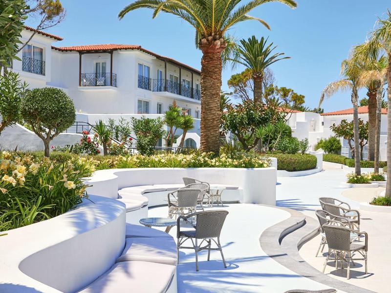 Moderní venkovní prostor s oblými bílými lavicemi, stoly a židlemi obklopený palmami.