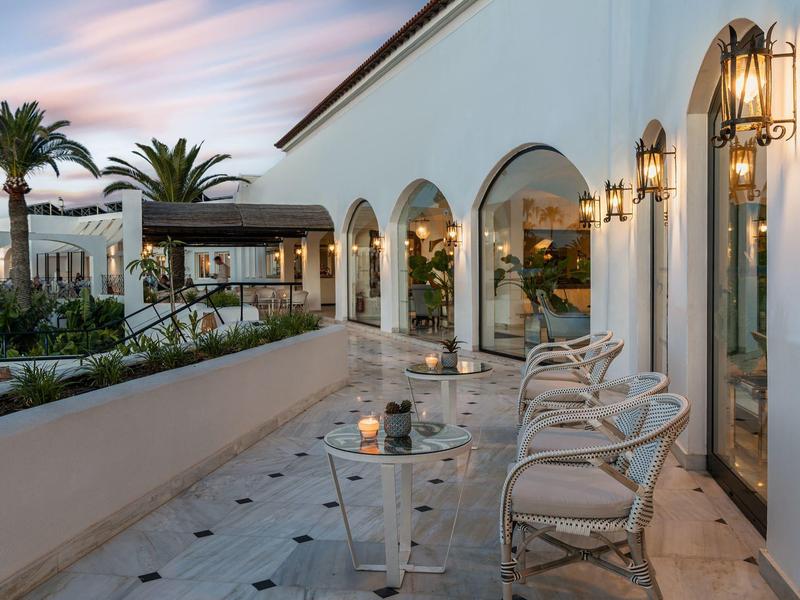 Terasa se židlemi a stolem vedle hotelu s dekorativními lucernami za západu slunce