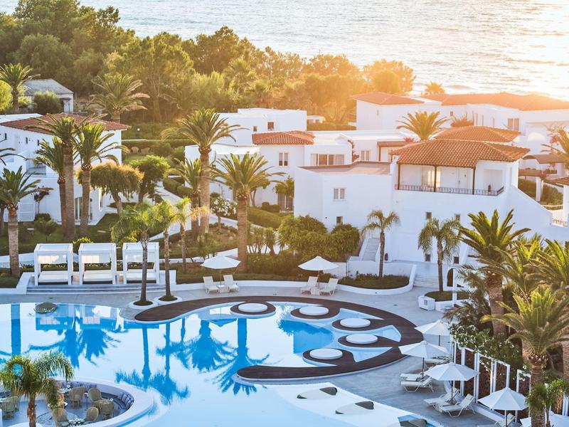 Bazén s bílými lehátky a palmami před bílými budovami u moře při západu slunce.