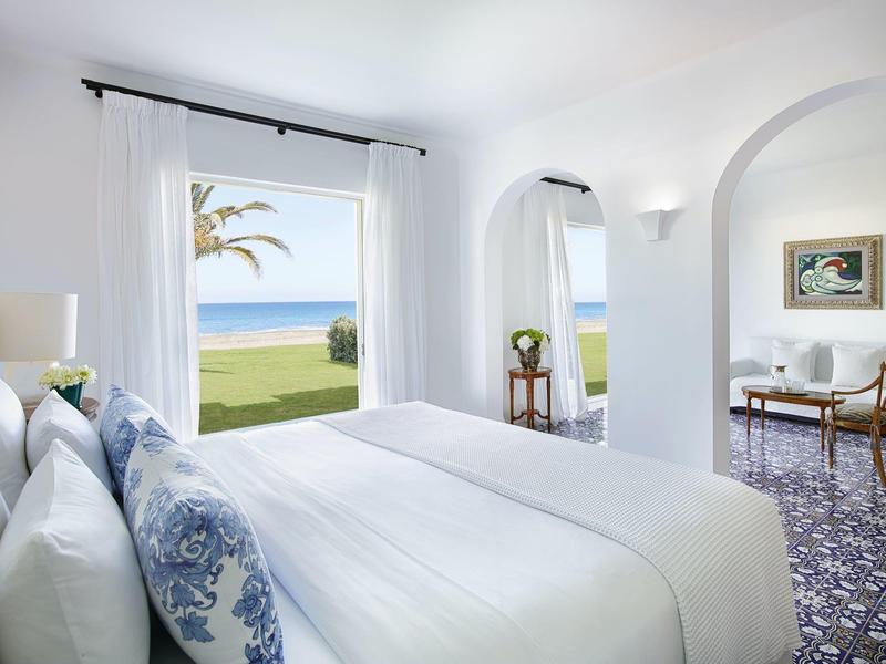 Světle ložnice s výhledem na moře, bílým povlečením a modro-bílou dlažbou.