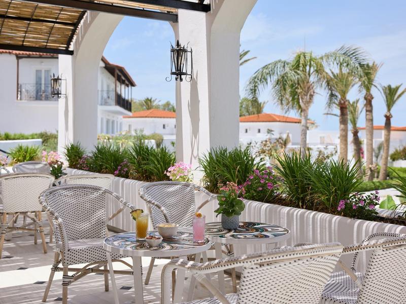 Moderní hotelová terasa s bílým nábytkem, rostlinami a výhledem na palmy za slunečného počasí.